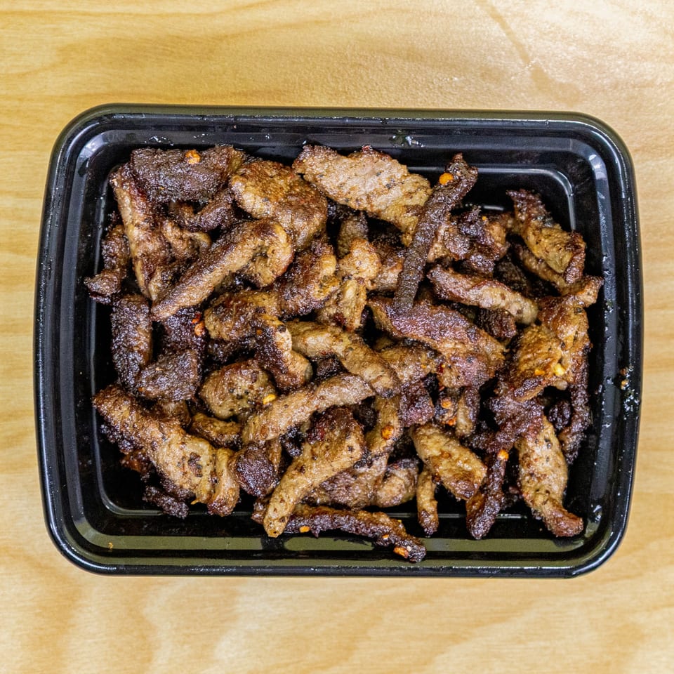A black takeout container filled with crispy, browned halal beef strips on a light wooden surface.