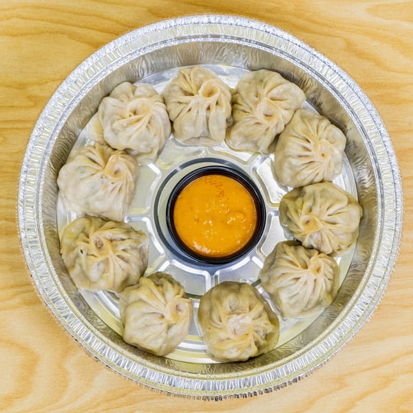 A dozen steamed halal momos arranged around a small cup of orange dipping sauce in a foil container.