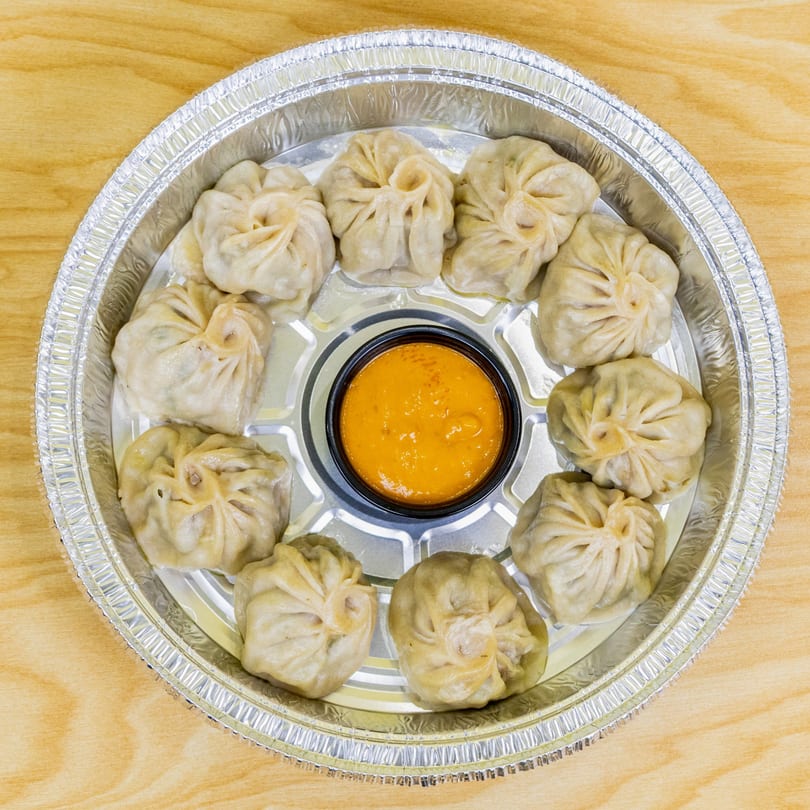 A dozen steamed halal momos arranged around a small cup of orange dipping sauce in a foil container.